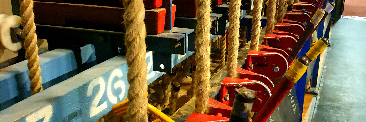 Close-up of a theatre fly system: a close-up of a series of thick, textured ropes descending vertically between blue and red structural supports. A glimpse of a floor with chalk markings can be seen.