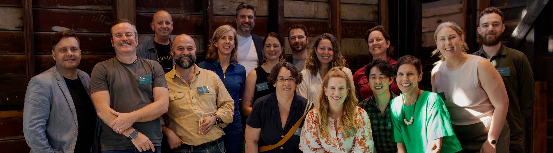 A group of sixteen diverse people - arts workers - are posed closely together, smiling and looking directly at the camera. They stand in front of a rustic wooden panel wall. Most are wearing name tags.