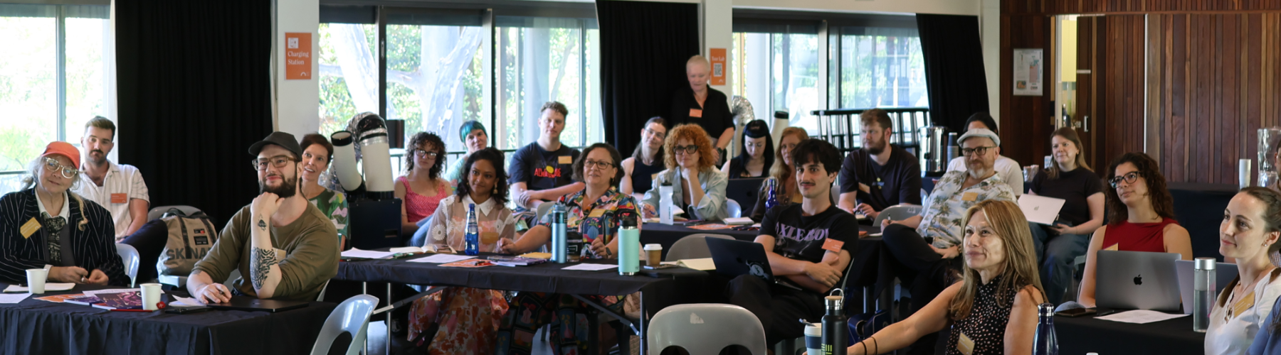 A group of people sit at tables in a workshop or presentation, facing forward.