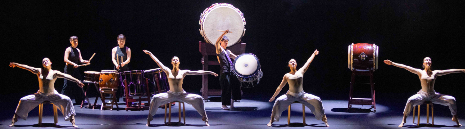 A panoramic stage view of a Taiko drumming and dance performance. Seven performers are visible: six are seated with outstretched arms. They are wearing neutral-toned leotards and loose white pants. Two performers behind them are playing taiko drums. 