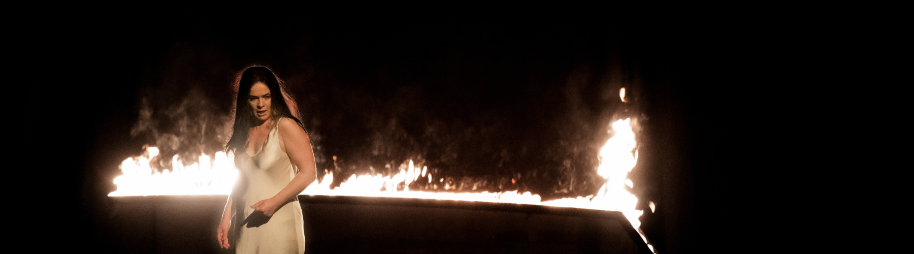 A woman in a pale slip dress stands in front of a wall of intense, bright flames in a dark setting.