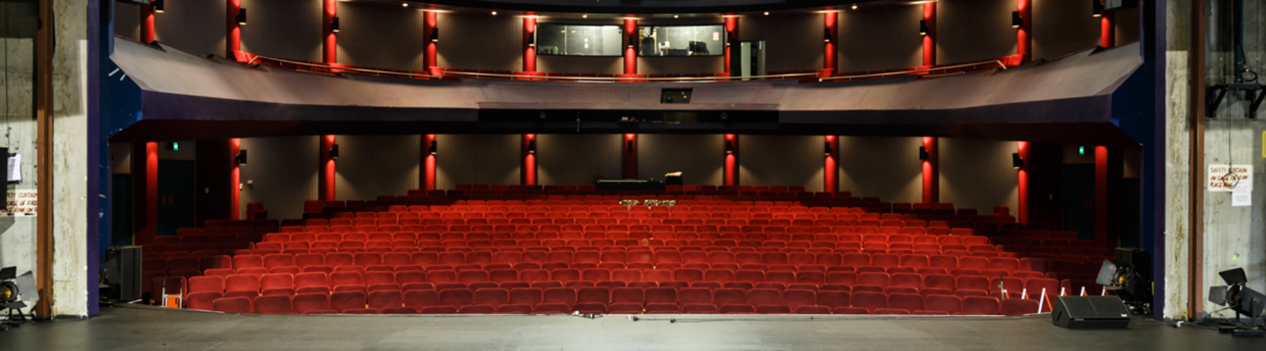 Panoramic view of an empty theater auditorium with rows of red velvet seats and a stage in the foreground.