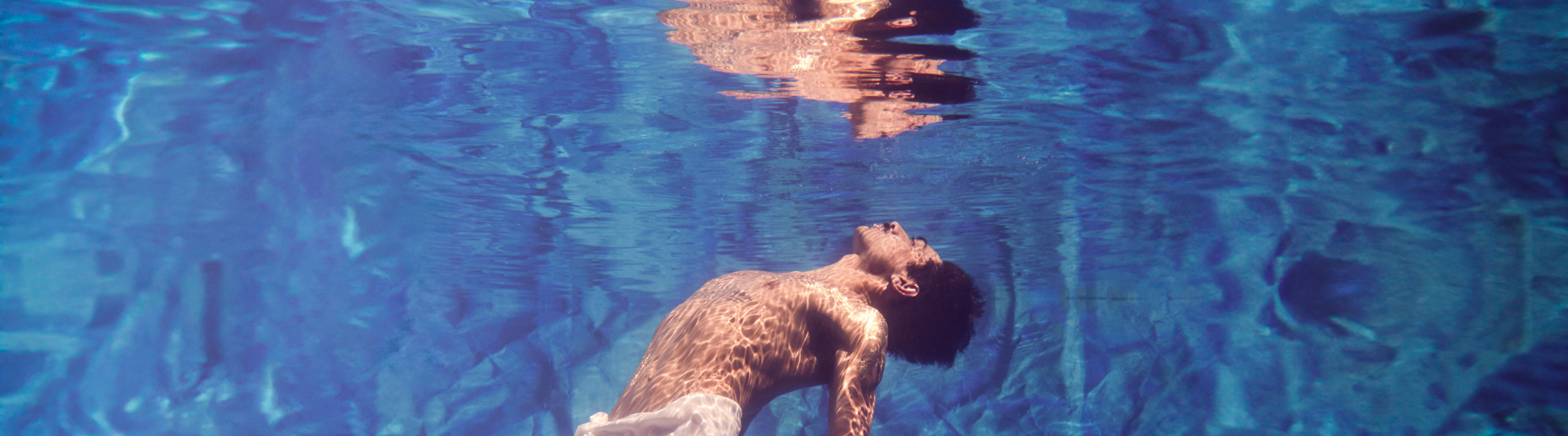 A man, shirtless and submerged in a body of blue water, looks upwards with his head tilted back; dappled sunlight appears on his torso and on the water's surface, where his reflection is visible.