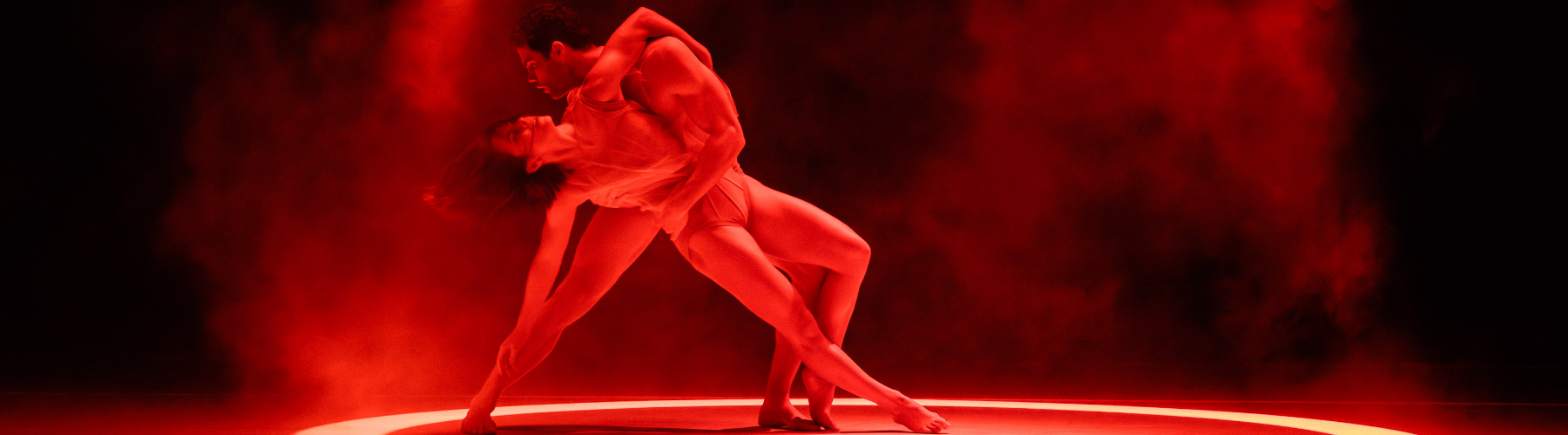 Two dancers in silhouette, bathed in red light and haze, performing a modern dance lift. The male dancer supports the female dancer's back as she leans backward, her body arched. The scene is lit with a bright ring of light at the base.