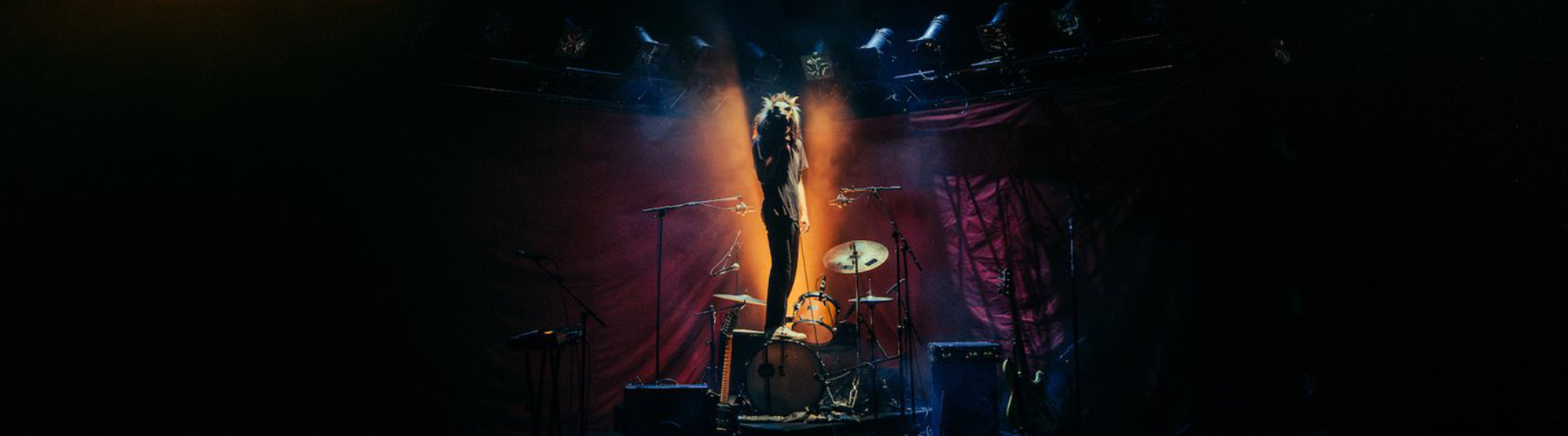Rock band instruments on a stage with dim lighting. A man with a wolf mask stands on top of the bass drum with a microphone in his hand.