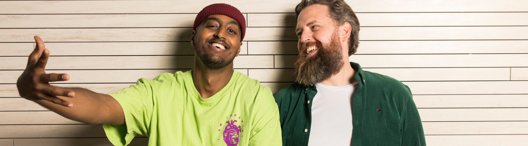 Two smiling men leaning against a wall. One is wearing a maroon beanie and lime green t-shirt, and has his right hand outstretched. The other man looks at the other and has his hands behind his back.