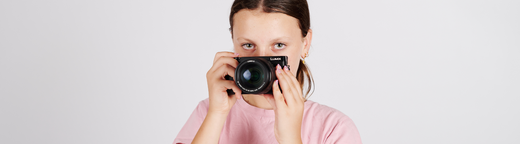 Young woman with brown hair in a pink shirt holds a black compact camera, looking directly at the viewer against a plain white background.