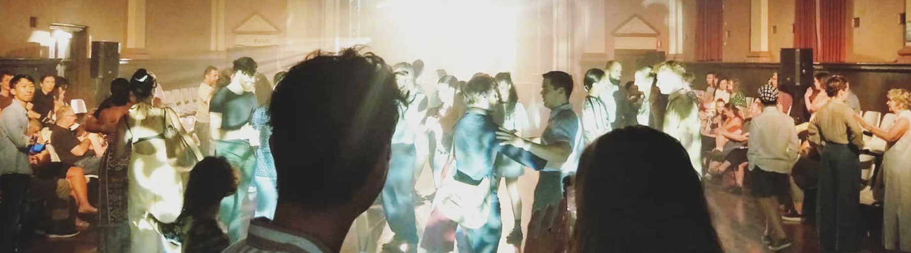 People dancing in a large, dimly lit town hall, with a bright light source visible in the distance, and the back of two people's heads in the foreground.