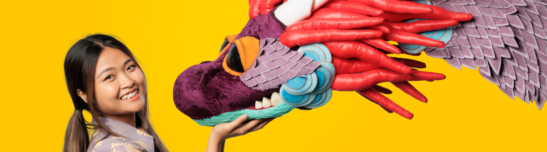 A young woman with dark hair smiling at the camera, with her hand on a colourful dragon puppet.