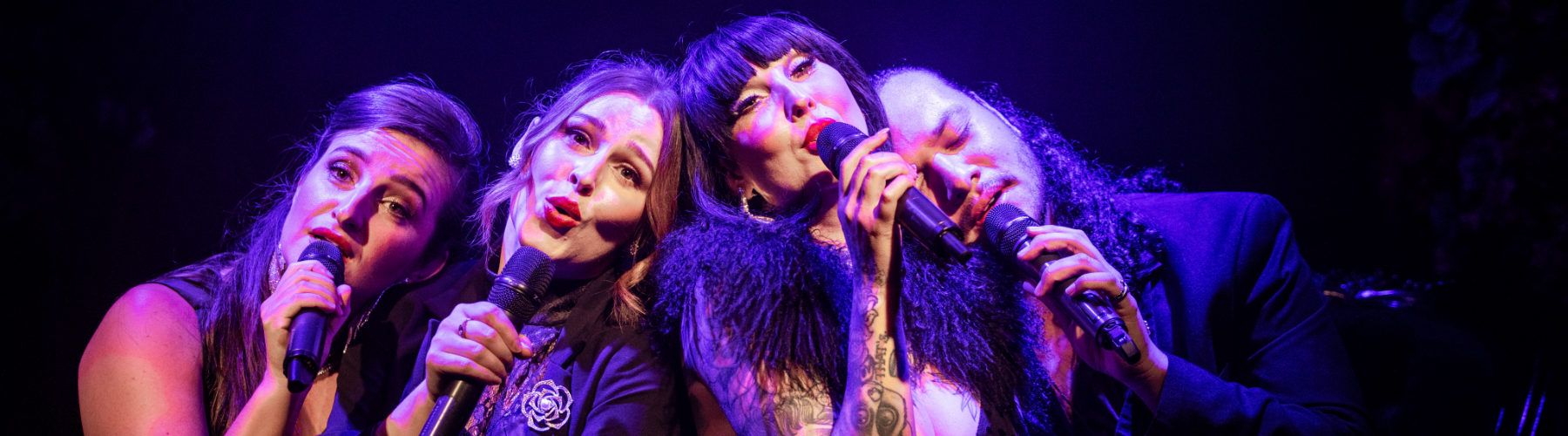 Four people singing into microphones on a dimly lit stage.