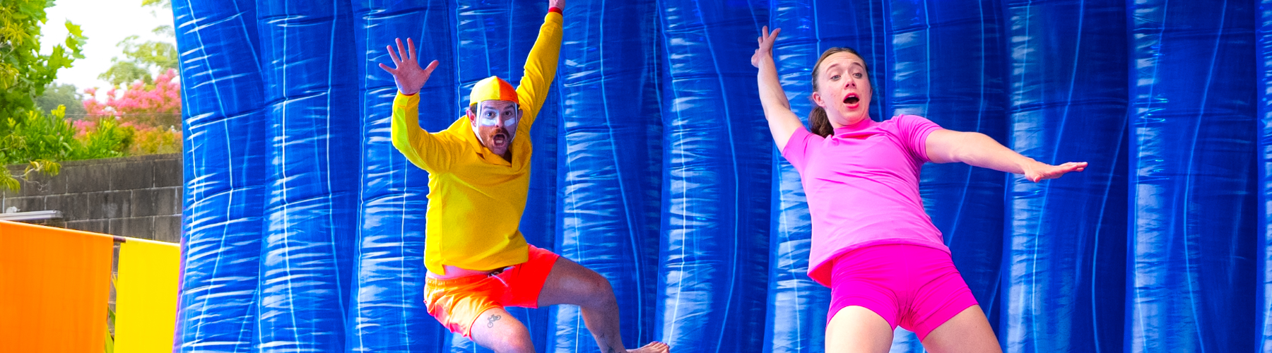 Two performers, one in yellow and red surf life saver attire, and the other in a pink swimwear, pretending to surf on an inflatable blue wave that is seen behind them.