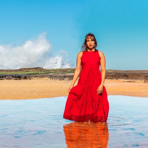 Woman in a red tiered dress stands knee-deep in calm water on a beach with a rock formation in the background and a large wave cresting in the distance under a bright blue sky.
