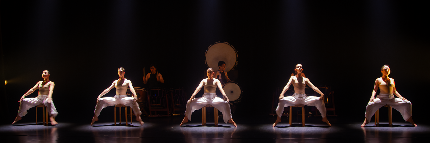 Five female dancers in white pants and tops, seated on wooden stools with legs wide apart, on a stage lit with spotlights. Behind them, two drummers play large, traditional drums. The backdrop is black.